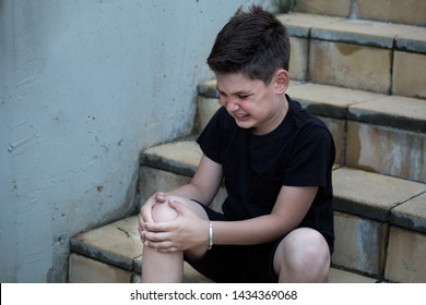 Boy In Pain Looking At His Injured Leg Sitting On The Stairs. Knee Pain