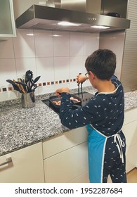 Boy In Pajamas And An Apron, Cooking Sausages In A Frying Pan In A Modern Kitchen, Preparing His Own Dinner, Vertical