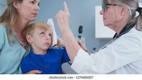 Boy At Pediatricians Office Holding Ice Pack On His Head With Mother And Doctor Checking On Concussion Symptoms. Child Using Cold Compress In Medical Exam Room Receiving First Aid