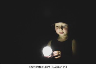 Boy pensive looking at burning light bulb dark black background low key portrait. Little child holding electric eco bulb hand thinking thought searching solution solving problem vintage cap retro