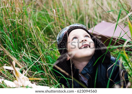 The boy in the pilot's helmet and glasses looks up at the sky, next to him is a toy airplane and a suitcase