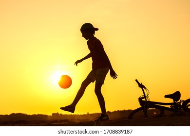 Boy playing with ball in nature, bicycle lies nearby, silhouette of playing child at sunset in countryside