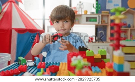 Boy playing with blocks at a table, hands touching colorful bricks and a toy car near a red tent in a building; imaginative curiosity.