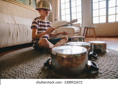 Boy playing drums on kitchenware at home. Boy pretending to be a drummer sitting on the living room floor and playing on utensils while wearing a bowl on the head.