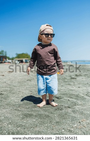 A boy is playing on the beach without shoes, wearing sunglasses and a cap. The sandy shore is bright under the clear sky. Waves are visible in the background.