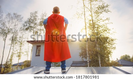 Boy is Playing a Role of a Super Hero. He's Standing on a Roof of a House with His Hands on His Waist. Young Man is Wearing a Bright Red Cape. He's Looking at the Sun.