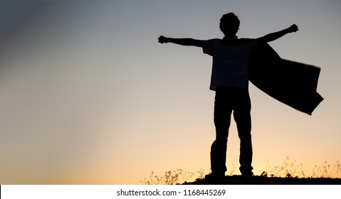 Boy Playing Superheroes On The Sky Background, Silhouette Of Teen Superhero In A Raincoat On The Hill