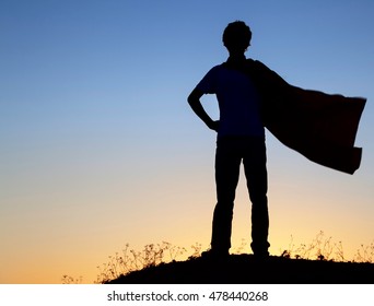 Boy Playing Superheroes On The Sky Background, Silhouette Of Teen Superhero In A Raincoat On The Hill