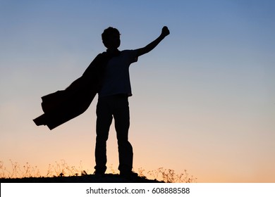 Boy playing superheroes on the sky background, silhouette of teenage superhero in a red cloak on a hill