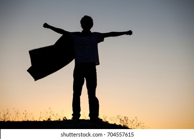 Boy Playing Superheroes On The Sky Background, Silhouette Of Teen Superhero In A Raincoat On The Hill