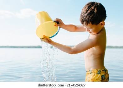 Boy Playing In Water In Nature. Child Pouring Water From Bucket While Standing In Lake Outdoors. Summer Vacation Concept.