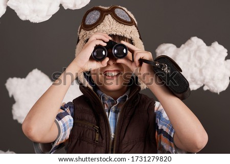 a boy plays with binoculars and dreams of becoming a pilot, dressed in a retro style jacket and helmet with glasses, clouds of cotton wool, gray background, tinted in brown