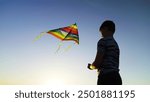 Boy plays kite with toy. Flying kite in blue sky, silhouette of child. Childhood, dream of flying, freedom travel. Happy boy playing with multi colored flying kite in nature in rays of summer sun.