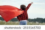 Boy plays superhero in red cape, childhood dream. Happy child playing superhero against sky. Little hero in red cloak watches sunset. Brave child winner in red raincoat plays in nature. Kid dreams