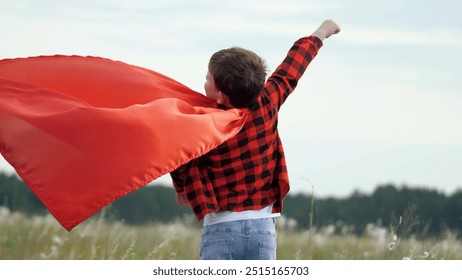 Boy plays superhero in red cape, childhood dream. Happy child playing superhero against sky. Little hero in red cloak watches sunset. Brave child winner in red raincoat plays in nature. Kid dreams
