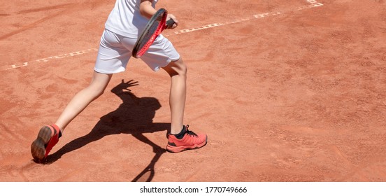 Boy plays tennis on red clay court. Child on tennis court hits forehand. Young tennis player with racket in action. Kids sports background with shadow. Banner size. Copy space