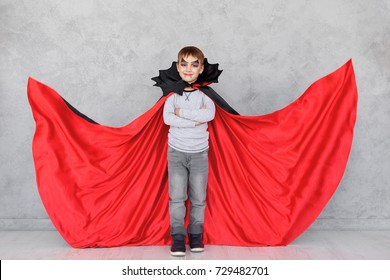 Boy  Portrait Wearing Halloween Vampire Makeup With Fluttering Red, Black Cloak, On A Gray Textured Background. Cute Kid Wearing Super Hero Costume With Crossed Hands, Copy Space Available.