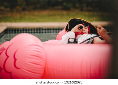 Boy pretending to be a pirate lying on inflatable mattress and looking through a binocular. Pirate looking with spyglass in swimming pool. - Powered by Shutterstock