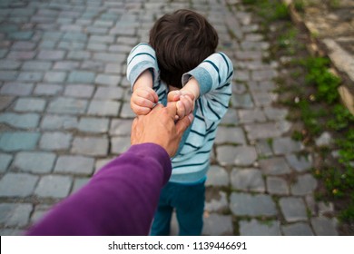 The Boy Is Pulling His Father's Hand. Dad Walks With His Son Along The City Street. The Child Is Capricious And Does Not Let Go Of The Parent's Hand.