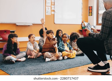 Boy raises his hand to answer a question in a classroom; he is sitting on the floor with other kids and the teacher is sitting in front of the class. Early child development in an elementary school. - Powered by Shutterstock