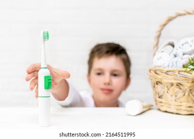 Boy Reaches Forward To Take The Electric Toothbrush And Start Brushing His Teeth In The Bathroom At Home. Dental Hygiene, Daily Morning Routine For Childrens Health. Caries Prevention And Protection.