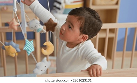 Boy reaches for a hanging mobile while standing and grasping crib rail in a nursery building; curiosity discovery play.