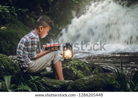 Boy reading book or bible with oil lamp at waterfall. Children and religion.