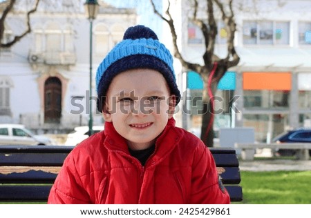 A boy in a red jacket and blue hat wrinkles his nose and bares his teeth. A six-year-old boy grimaces, showing an angry face.