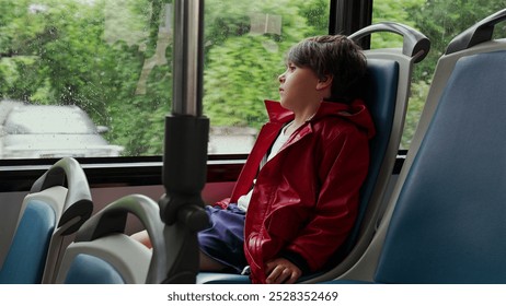 Boy in a red jacket gazing thoughtfully out a bus window on a rainy day, with empty blue seats around him. reflective mood as the wet streets blur outside the window - Powered by Shutterstock