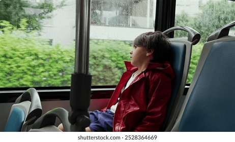 Boy in a red jacket gazing thoughtfully out a bus window on a rainy day, with empty blue seats around him. reflective mood as the wet streets blur outside the window - Powered by Shutterstock