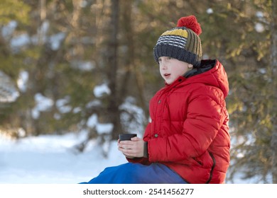 Boy in red jacket and hat sitting on the snow with a cup in his hand, sits and looks into the distance.