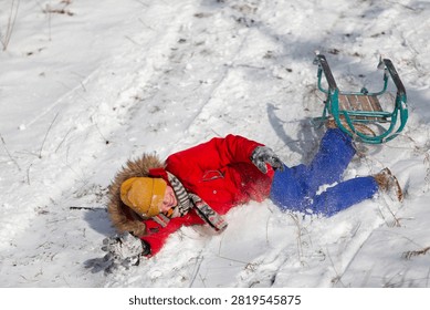 A boy in a red jacket, yellow hat and blue pants playfully falls from a green sled on a snowy slope. The sun casts shadows on the white snow, and a scarf trails behind it.