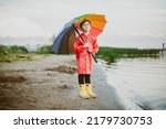 Boy in a red raincoat and yellow rubber boots stands at river bank and holding rainbow umbrella. School kid standing still near autumn lake. Child wearing waterproof clothes at shoreside.
