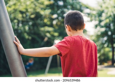 Boy with red t-shirt stands leaning against a metal post and watches as a child rides on the cable car in the park 