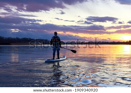 Boy rowing on stand up paddle boarding (SUP) paddling along the calm autumn Danube river against background of a colorful sunset. The concept of children's sports and tourism.