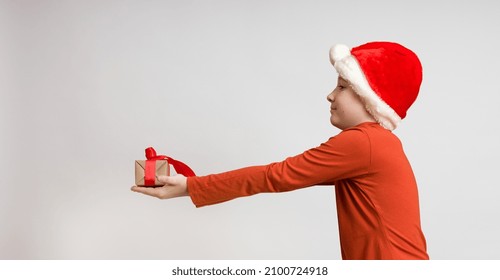 Boy In Santa Hat Gives A Gift In Outstretched Hands On A White Background, Side View