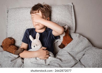 The boy was scared before going to bed. Night terrors in a child. The kid covers his face with his hands in a fear. Children's experiences. Boy in the bed. View from above. Sad psychological state.