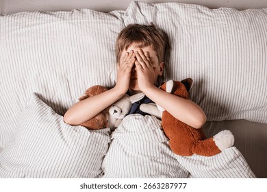 The boy was scared before going to bed. Night terrors in a child. The kid covers his face with his hands in a fear. Children's experiences. Boy in the bed. Air alarm. Sad psychological state.
