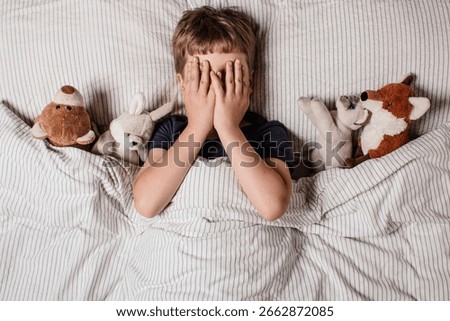 The boy was scared before going to bed. Night terrors in a child. The kid covers his face with his hands in a fear. Children's experiences. Boy in the bed. View from above. Sad psychological state.