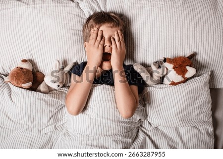 The boy was scared before going to bed. Night terrors in a child. The kid covers his face with his hands in a fear. Children's experiences. Boy in the bed. Air alarm. Sad psychological state.