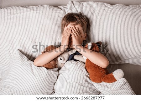 The boy was scared before going to bed. Night terrors in a child. The kid covers his face with his hands in a fear. Children's experiences. Boy in the bed. Air alarm. Sad psychological state.