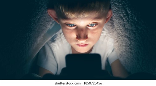 Boy with a serious face under the blanket at night in his bed communicates on Internet. Child gadget addiction and insomnia. - Shutterstock ID 1850725456