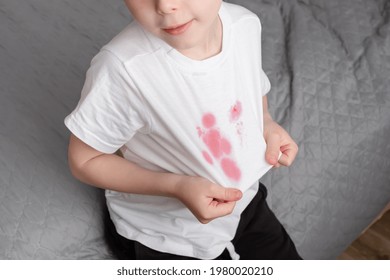 Boy Showing A Stain Spilled Juice On His White T-shirt, Sitting On The Bed. The Concept Of Cleaning Stains On Clothes. 