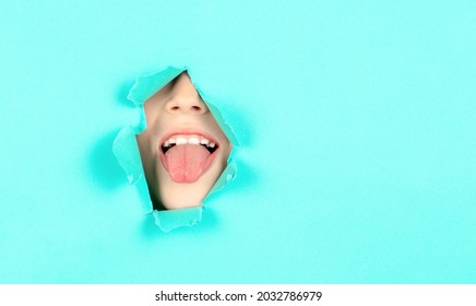 Boy shows his tongue through hole in the wall. Portrait funny kid man showing tongue in a torn paper hole. Close-up Boy Face Showing Her Tongue. Child stuck out her tongue through a hole in the paper.