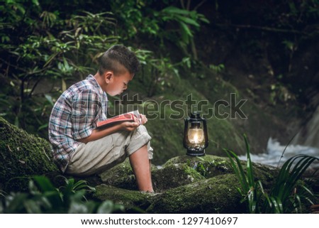 Boy siting on green rock and reading book or bible with oil lamp at waterfall. Children and religion.