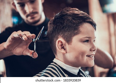 A boy sits in a barbershop while he is being cut with scissors. Little boy smile while hairdresser cuts his hair with scissors.  - Powered by Shutterstock