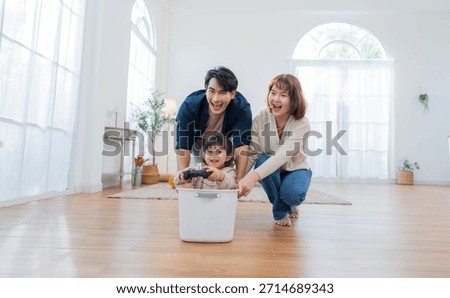 A boy sits in a box while his parents push him around the living room. The room has large windows and a wooden floor. Laughter fills the air during their playtime.