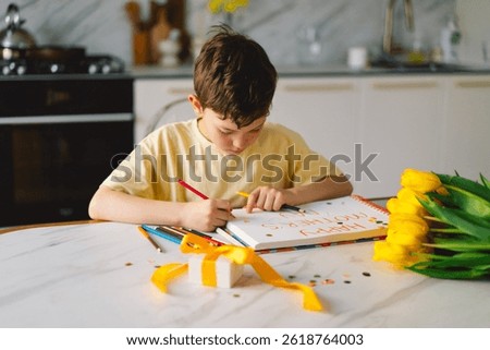 A boy sits at a kitchen table, diligently creating a heartfelt Mother's Day card, with the message Happy mothers day. Beautiful yellow tulips and colorful pencils are scattered around.