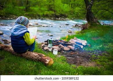 The boy sits on the Bank of the river near the bonfire and holds the map, near the fire is a mug and a coffee maker