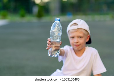 A Boy Sits On A Green Lawn On A Football Field And Holds A Bottle Of Water. Focus On The Water Bottle.